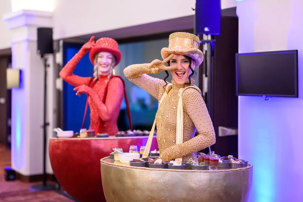 Zwei Performerinnen in Rot und Gold mit Zylinderhüten begrüßen die Gäste